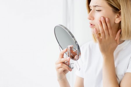 Eine Frau begutachtet ihre zu Akne neigende Haut im Spiegel, zufrieden mit der Verbesserung  durch die Befolgung der Routine für reine Haut.